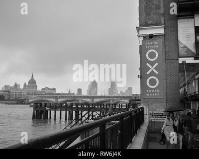 B&W immagine di due signore vestito per una notte fuori Londra, passando la OXO Tower sulla riva sud del Tamigi, ombrelli azienda contro la pioggia. Foto Stock