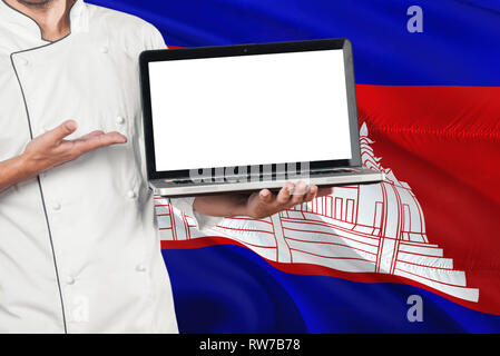 Lo Chef cambogiano holding portatile con schermo vuoto sulla Cambogia bandiera dello sfondo. Cuocere indossano uniformi e laptop di puntamento per copiare lo spazio. Foto Stock