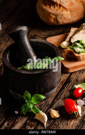 Mortaio con pesto genovese. Sfondo di legno con vari tipi di pane, basilico, aglio, pomodori e cucchiaio Foto Stock