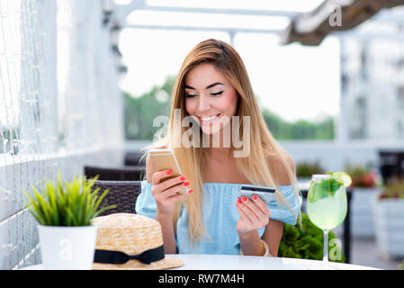 Giovane e bella ragazza sorridenti in estate cafe restaurant. In mano il telefono di plastica e carta di credito. Applicazione di online banking online Foto Stock