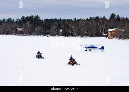 Due motoslitte attraversando il Lago Pleasant nelle Montagne Adirondack, NY USA passato un Cub Crafters velivoli sperimentali su sci parcheggiata sul ghiaccio. Foto Stock