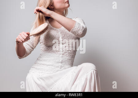 Giovane donna in bianco spazzolando i suoi lunghi capelli biondi con una spazzola in legno. Foto Stock