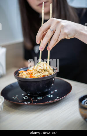 Stretta di mano umana grabbing tagliatelle con bacchette da un recipiente con acqua calda zuppa di noodle. Foto Stock