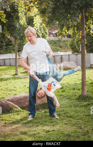 Padre Felice con il suo piccolo figlio in parco nella giornata di sole. Papà detiene il figlio a testa in giù Foto Stock