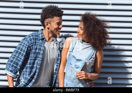 Giovane ragazzo e ragazza isolato permanente sulla parete avvolgente guardando ogni altro ridere giocoso Foto Stock