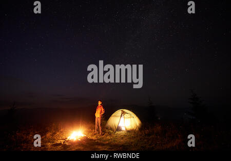 Notte in campeggio in montagna. Giovane ragazza turistica in piedi vicino alla tenda illuminata e bruciare il fuoco su deep dark sky con sacco di luminose stelle scintillanti sullo sfondo. Turismo e attività outdoor concept. Foto Stock