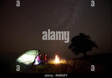 Turista giovane uomo e donna avente un resto vicino alla tenda illuminata, bruciando il fuoco e la silhouette di un grande albero sotto il cielo stellato, godendo di notte campeggio in montagna. Turismo, attività outdoor concept Foto Stock