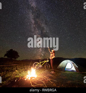 Giovane donna di riposo turistico di notte campeggio, puntando alla incredibile bella serata cielo pieno di stelle e la Via Lattea, in piedi vicino al fuoco ardente, tenda illuminata, mountain bike. Il concetto di turismo Foto Stock