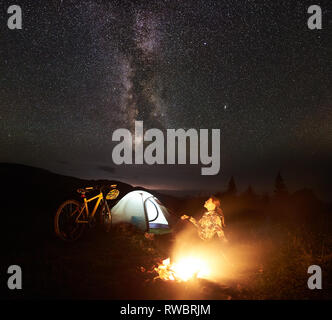 Giovane donna tourist godendo di notte campeggio vicino a bruciare falò, illuminato tenda turistica, mountain bicycle sotto incredibile bella serata cielo pieno di stelle e la Via Lattea. La fotografia astronomica Foto Stock