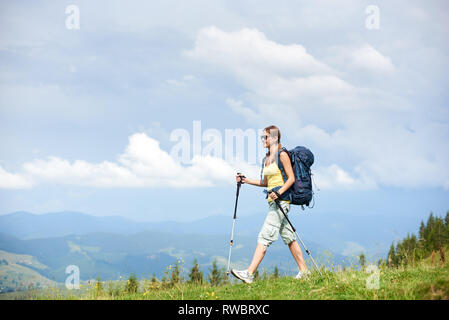 Donna sorridente escursionista trekking in montagna dei Carpazi trail, camminando sulla collina erbosa, indossando uno zaino e occhiali da sole, utilizzando bastoncini da trekking, godendo di estate giorno nuvoloso. Attività all'aperto, il concetto di turismo Foto Stock