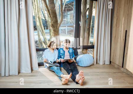 Felice coppia seduti insieme sul pavimento vicino alla finestra con splendida vista, la lettura di alcune riviste e un momento di relax a casa Foto Stock