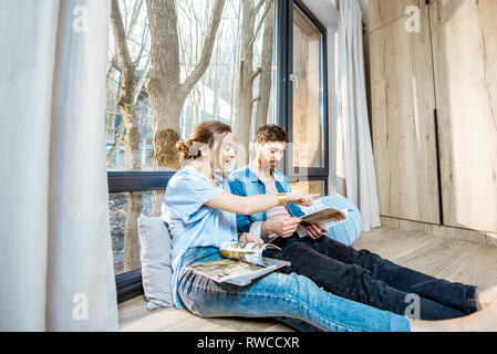 Felice coppia seduti insieme sul pavimento vicino alla finestra con splendida vista, la lettura di alcune riviste e un momento di relax a casa Foto Stock