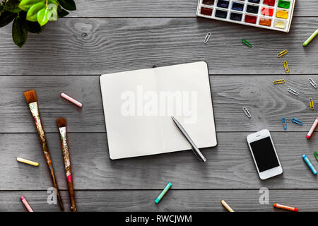 Colorate artista creativa tabella vuota con il notebook per lavorare i bozzetti e vernici, arte pennelli forniture impostato sul grigio Sfondo di legno, vista dall'alto, co Foto Stock