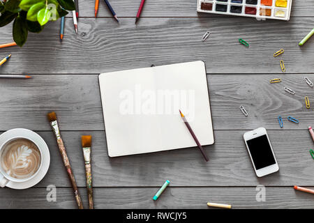 Colorate artista creativa tabella con blank notebook vernici a matita per schizzi di lavoro, arte pennelli forniture impostato sul grigio Sfondo di legno, vista dall'alto, Foto Stock