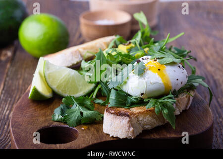 Sandwich vegetariano con uovo in camicia, avocado, rucola su fette di pane tostato e sana colazione o uno spuntino rustico di legno, fuoco selettivo, chiudi Foto Stock