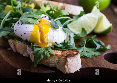 Sandwich vegetariano con uovo in camicia, avocado, rucola su fette di pane tostato e sana colazione o uno spuntino rustico di legno, fuoco selettivo, chiudi Foto Stock