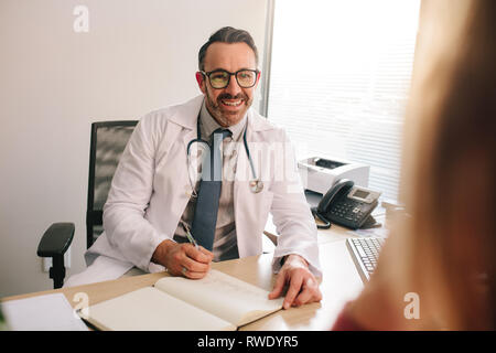 Medico di sesso maschile seduto alla sua scrivania di avvalersi della consulenza di un paziente. Medico di medicina generale a parlare con il paziente nella sua clinica. Foto Stock