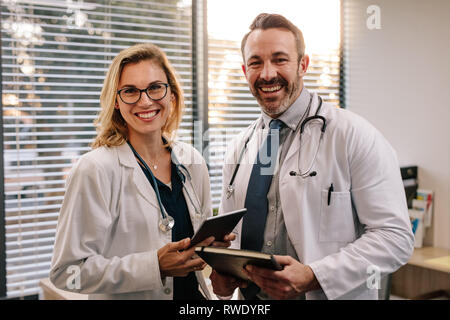 Ritratto di due medici sorridente e guardando la fotocamera mentre si tiene un tablet e un diario in clinica. Due medici sorridente in camice bianco workin Foto Stock