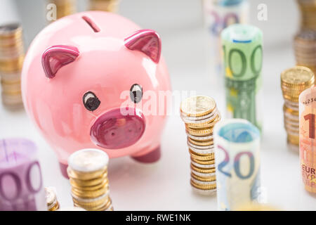 Rosa salvadanaio in medio dei rotoli di banconote in euro e torri con le monete Foto Stock