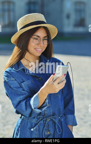 Un attraente giovane donna asiatica indossando occhiali da sole e una tuta in denim in piedi fuori con un color argento telefono Foto Stock