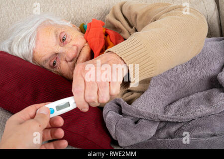 Close up foto donna senior essendo malato, accompagnatore tenendo il termometro digitale accanto alla sua testa - febbre Foto Stock