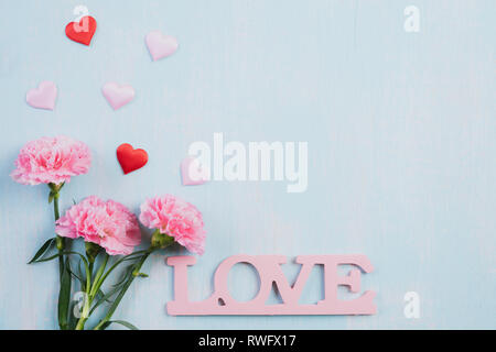 Il giorno di San Valentino e il concetto di amore. Rosa fiori di garofano, legno lettere che compongono la parola amore scritta con cuore rosso blu su sfondo color pastello. Foto Stock