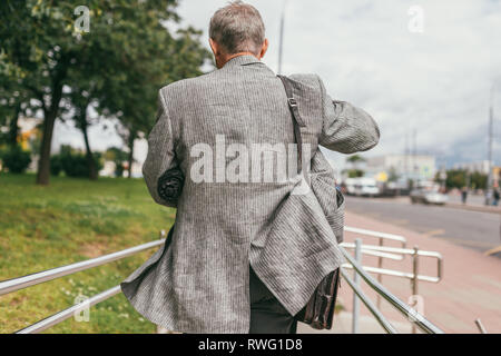 Un capelli grigi uomo anziano in una giacca grigia con un ombrello sotto il suo braccio e una pelle marrone a sacchetto sulla sua spalla correndo giù per la strada. Vista posteriore Foto Stock