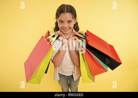 Esperto di moda. Bambino graziosi negozi expert aiuta a portare pacchetti durante lo shopping. Little Shop esperto. Shopping ragazza ama fare shopping. Kid ragazza faccia felice porta mazzetto pacchetti sfondo giallo. Foto Stock