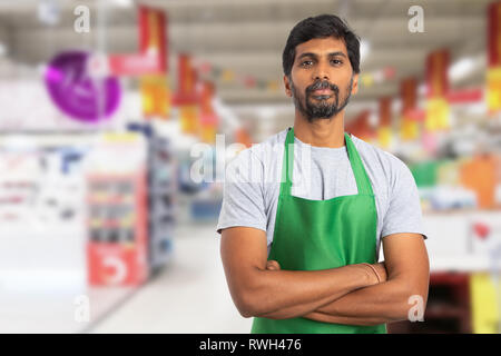 Maschio indiano il supermercato o ipermercato dipendente ritratto foto indossando grembiule verde con bracci incrociati e espressione seria Foto Stock