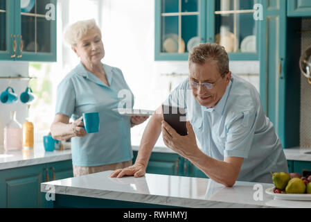 Uomo in pensione, addicted allo smartphone, la lettura di notizie, mentre sua moglie arrabbiato portò una tazza di tè. Dipendenza da digitale a rovinare la vita. Foto Stock