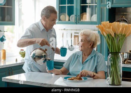 L'uomo versando il tè in moglie per la tazza. Coppia di anziani aventi un buon tempo insieme in cucina. Giornata della donna concetto. Foto Stock