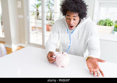 African American uomo con uno stetoscopio controllo salute finanziaria sul porcellino salvadanaio paura in stato di shock con una sorpresa faccia paura ed eccitati con timore ex Foto Stock