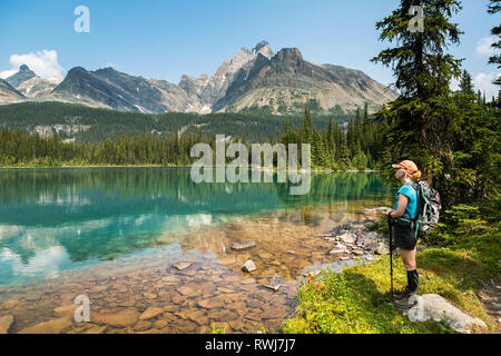 Escursionista femmina in piedi lungo la riva di un lago alpino refelcting mountain range con cielo blu; British Columbia, Canada Foto Stock