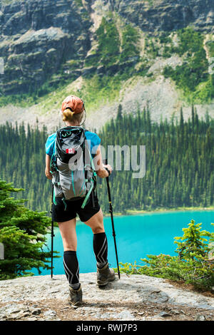 Escursionista femmina permanente sulla scogliera rocciosa che si affaccia colorato lago alpino e la scogliera di montagna in background; British Columbia, Canada Foto Stock