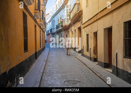 Un anziano uomo cammina giù per la strada stretta tra edifici residenziali; Cadice, Andalusia, Spagna Foto Stock