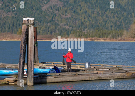 Donna di mezza età in bright cappotto invernale in piedi su un molo con kayak pagaie. Concessione si restringe il Parco Regionale, Pitt Meadows, B.C., Canada Foto Stock