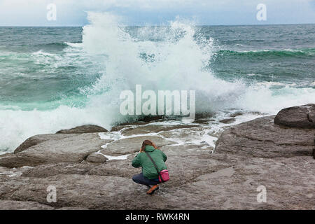 Giovane uomo accovacciato su una sporgenza di roccia nel tentativo di fotografare le onde del mare da vicino. Port au choix National Historic Site, Port au choix, Terranova, Canada Foto Stock