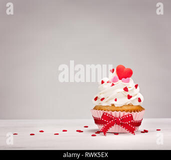 Cupcake alla vaniglia con glassa di crema di burro, a forma di cuore e spruzza un fiocco rosso. Foto Stock