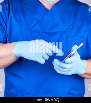 Dottore in uniforme blu e guanti in lattice possiede un telefono cellulare e tocca lo schermo Foto Stock