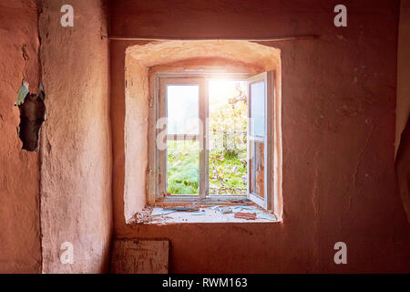 Interno di una rovina il vecchio cottage con una luce rosa di parete e la rottura di una finestra in legno telaio guardando attraverso un rurale prato verde paesaggio del campo Foto Stock