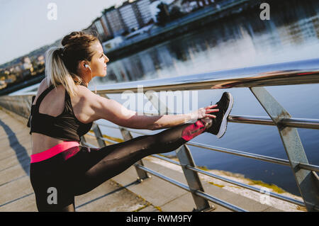 Signora giovane in abbigliamento sportivo gambe di stiro sul molo vicino acqua in città nel giorno di sole Foto Stock