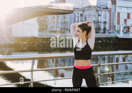 Signora giovane in sportswear stretching mani sul molo vicino acqua in città nel giorno di sole Foto Stock