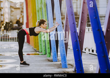 Vista laterale della giovane donna in abbigliamento sportivo con gli auricolari e lo smartphone stretching gambe su strada Foto Stock