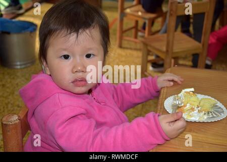 Il cantone, Cina - CIRCA NEL MARZO 2019: adorabile bambina di mangiare una fetta di torta in una scuola materna. Foto Stock