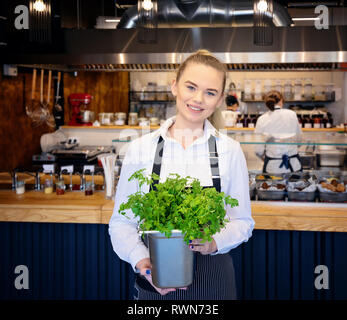 Sorridente giovane donna holding di prezzemolo fresco. Ristorante sfocato il contatore e i dipendenti che lavorano in background Foto Stock