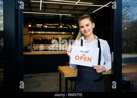 Portrait of a happy waitress standing at restaurant entrance. Portrait of young waitress attend new customers. Woman showing open sign at restaurant d Foto Stock