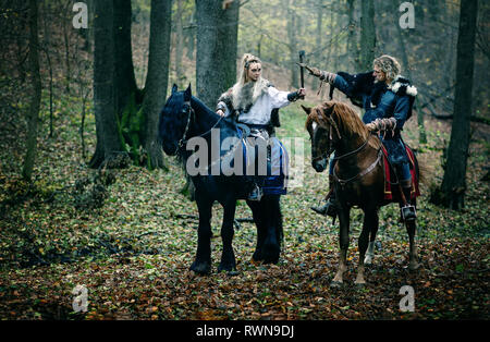 Guerrieri donna un uomo a cavallo nei boschi. Vichinghi scandinavi di equitazione con assi in mani. Abiti tradizionali con collare in pelliccia. Reconstructio Foto Stock