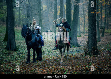 Guerrieri donna un uomo a cavallo nei boschi. Vichinghi scandinavi di equitazione con assi in mani. Abiti tradizionali con collare in pelliccia. Reconstructio Foto Stock