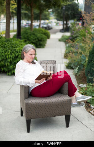 Una donna matura si siede a leggere un libro in una poltrona su una passerella esterna; Bothell, Washington, Stati Uniti d'America Foto Stock