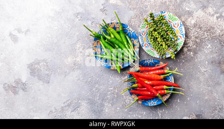 Peperoni di vari tipi di rosso verde nero in una ciotola con ornamenti colorati su sfondo grigio. Schema piatto. Copia dello spazio. Vista da sopra. Foto Stock